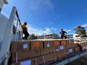 Two women stand on top of a truck facing the camera. The truck is filled with cardboard boxes, and is outside on the street. There are blue skies overhead.