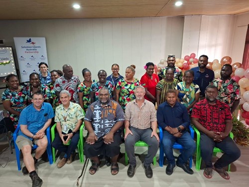 20 people pose for a photo in 2 lines, in the World Scabies Program Office in Honiara, Solomon Islands. 6 men sit on chairs in the front, and the rest of the people stand behind them.