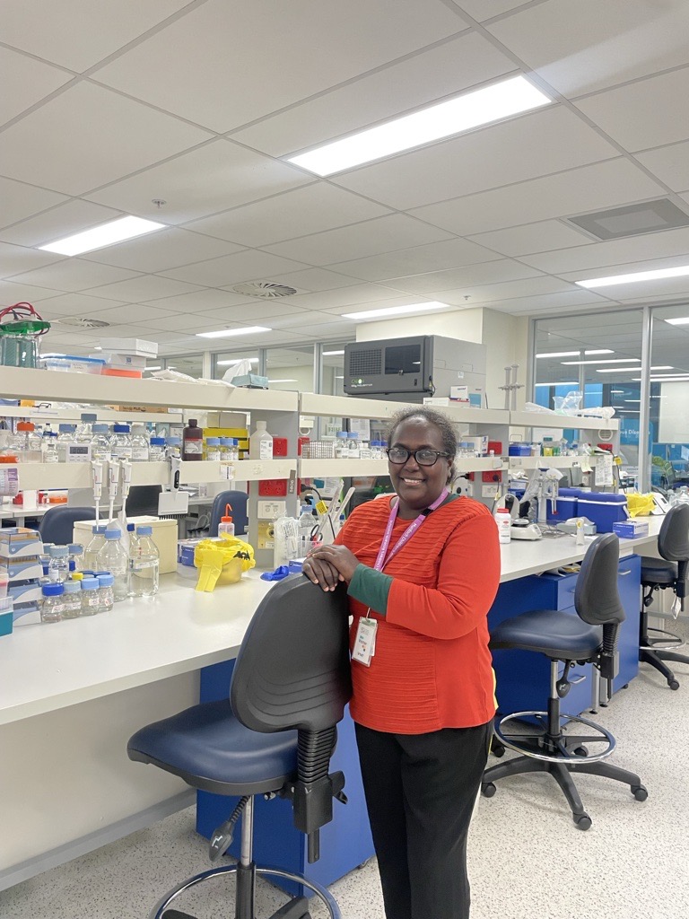 A woman standing in a laboratory, smiling at the camera. She is wearing glasses, an orange-red long-sleeve top with green sleeves underneath, black pants, and a purple lanyard with an ID badge. She is leaning on the backrest of a black laboratory chair. T