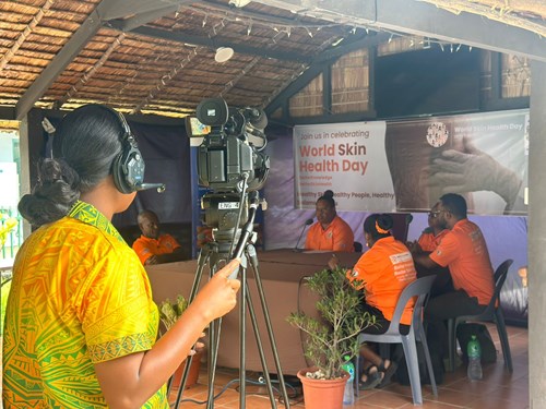 5 people sit around a table for a talk back radio show with SIBC news Solomon Islands, in Honiara on July 8 as part of World Skin Health Day. A woman is standing in the foreground ready to film them, she is wearing a headset.