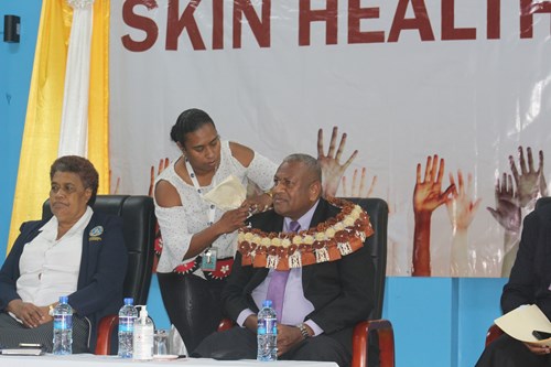 A woman assists a seated man wearing a ceremonial garland by adjusting a microphone on his collar during a World Skin Health Day event. Another woman sits beside him, and a backdrop with raised hand graphics and large text is visible behind them.