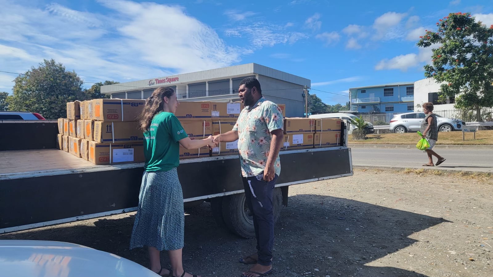 A man and a woman stand on the street in front of a truck that is being filled with cardboard boxes. They are facing each other shaking hands.