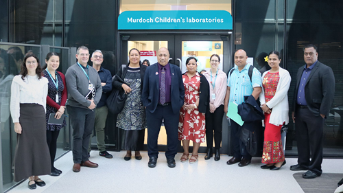 11 people stand in front of a glass door. Above the door is a sign with Murdoch Children's laboratories. Everyone is smiling.
