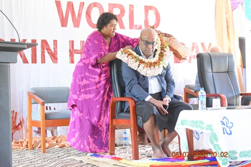 A woman with a pink bula dress in placing a garland around a man who is sitting down. He is wearing a suit jacket and glasses. They are on a stage with a World Skin Health Day sign behind them.