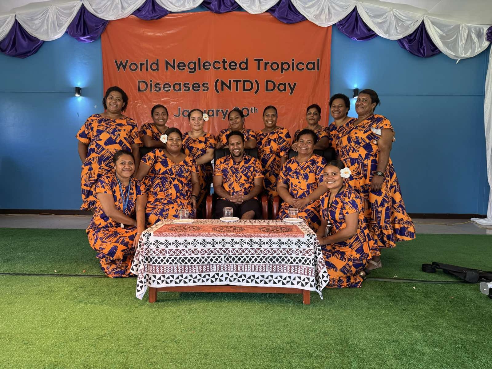 A group of people in matching orange dresses and t-shirts are around a table with a sign in the background for world NTD day