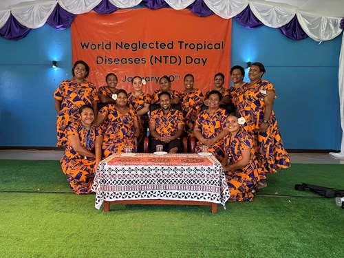 A group of people in matching orange dresses and t-shirts are around a table with a sign in the background for world NTD day