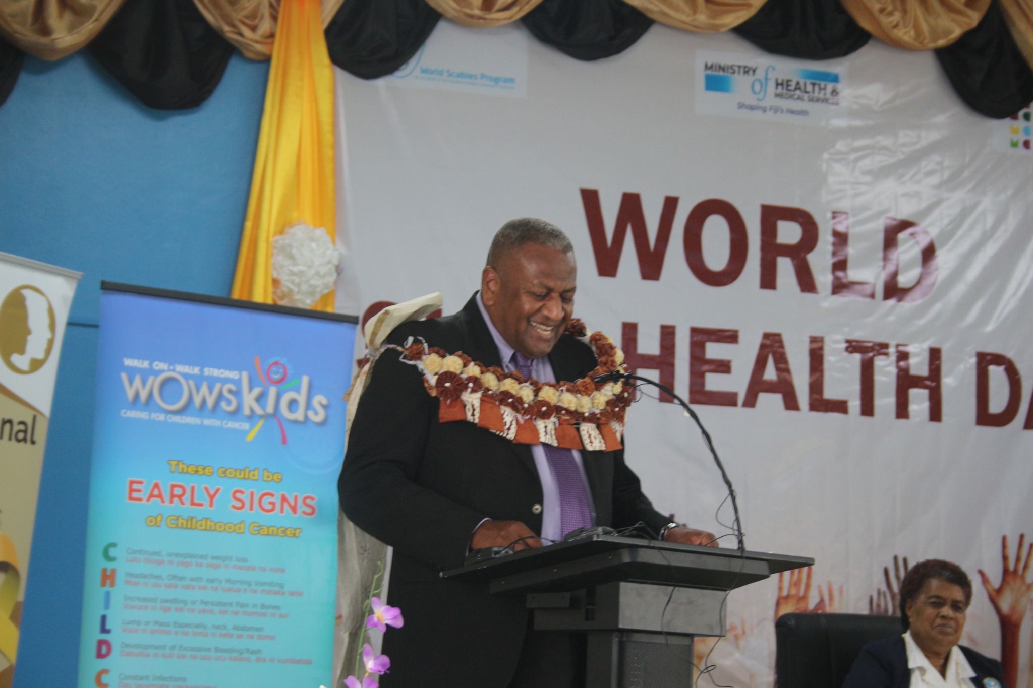 A smiling speaker in a suit and traditional Fijian garland stands at a podium during a World Health Day event. Behind him is a banner that reads "World Health Day" and a poster promoting early signs of childhood cancer by WOWS Kids Fiji. Another attendee