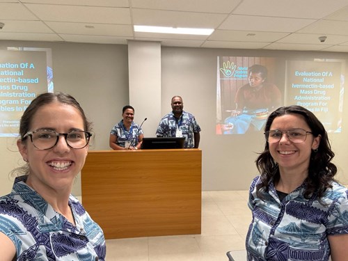 4 people pose for a selfie with matching Bula shirts in a conference room. 2 are women in close range on either side of the image, and there are two people (woman on the left, man on the right) in the centre who are further back and standing behind a podium. There is the title slide of a presentation on the wall of the room.