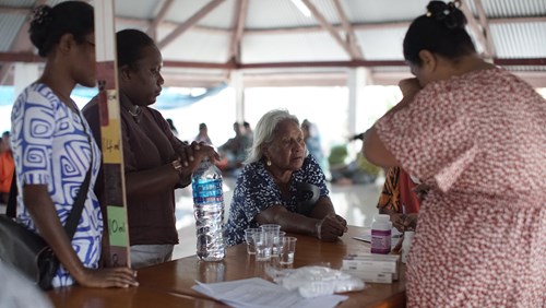 An elderly woman with white hair sits at a wooden table inside a community hall, engaging with a group of women providing healthcare support. Several plastic cups, a water bottle, medicine bottles, and paperwork are visible on the table. The setting appears informal and supportive, with the group focused on delivering medical treatment and information.