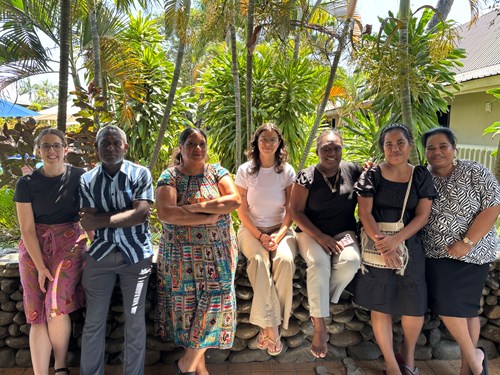 7 people Stand side by side looking at the camera for a photo. They are in a tropical place, with palm trees in the background. There is a ledge made of rocks which some people are sitting on and the others are leaning on.