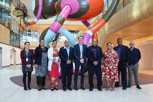 9 people are standing in the foyer of the Royal Children's Hospital. They are standing in front of a big art installation that looks like a colourful bug. The bug has a colourful butterfly on is. They are all smiling.