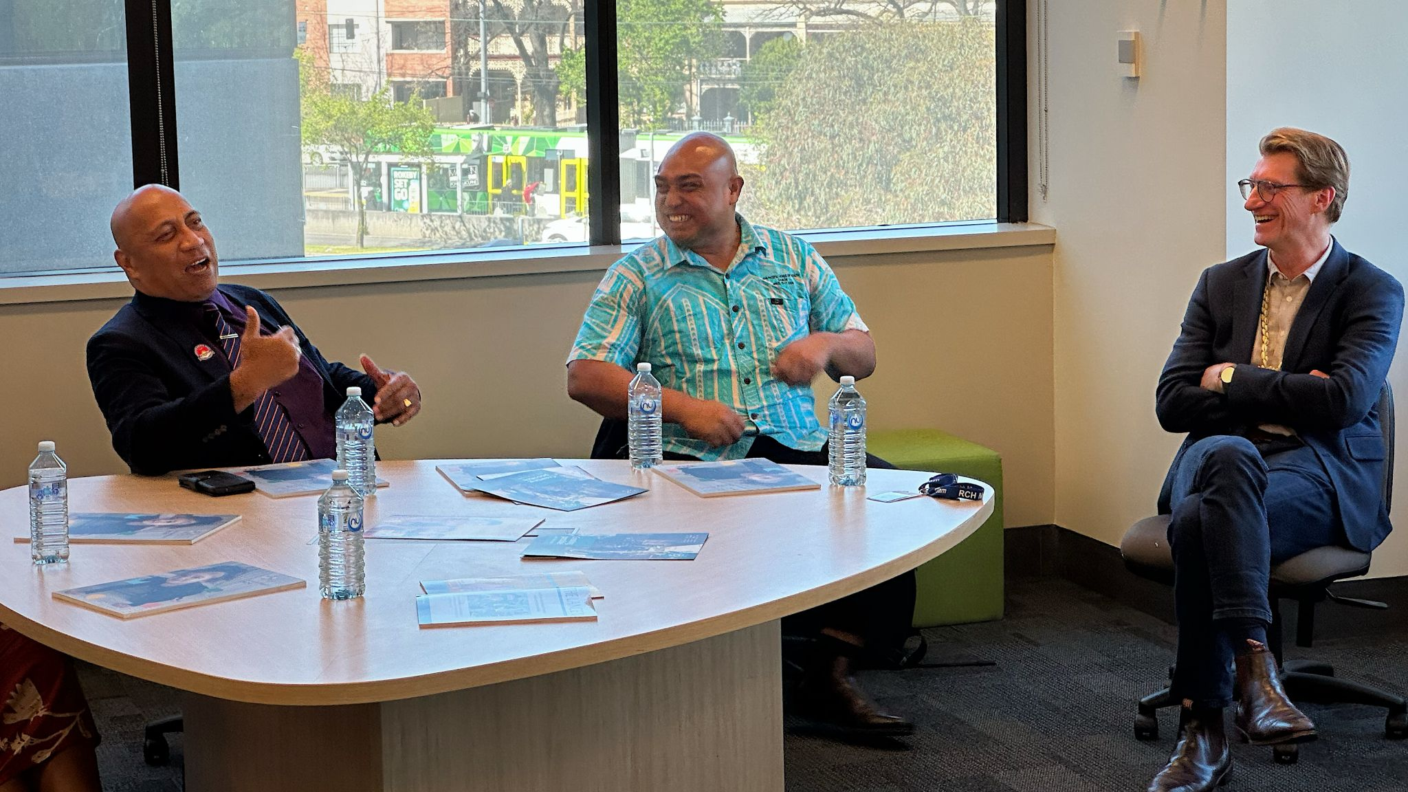 3 men are sitting around a table with a window in the background. One person is talk and the other two are laughing. There is a tram outside the window and some trees. There are 5 bottles of water and some flyers on the table.