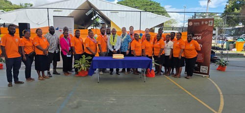 20 people stand in a row, posing for a photo to celebrate the World Skin Health Day launch event on July 8 in Honiara, Solomon Islands. There is a table in the centre with a cake on it.