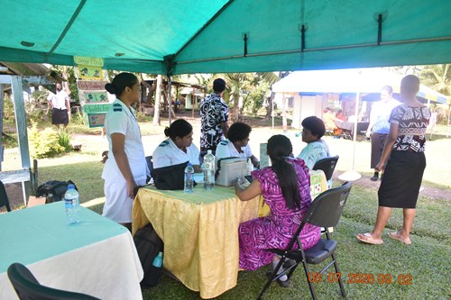 A group of people are sitting around a table under a green tent in a community setting. There are three nurses in white uniforms, one person it a bright pink bula dress and a community member receiving a skin screening