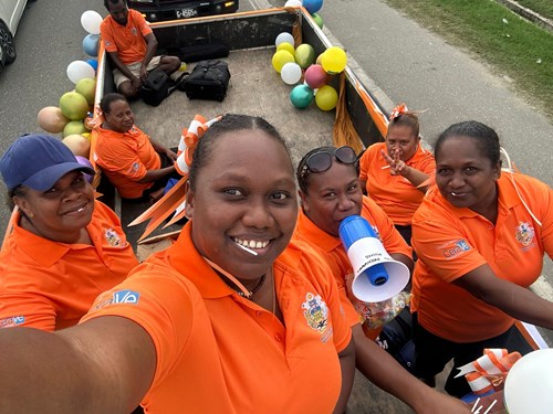 5 women pose for a selfie in a truck. 2 others, a man and a woman, are sitting in the background in the truck. They are all wearing orange T shirts in celebration of world skin health day, on the 8th of July in Honiara, Solomon Islands.