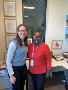 Two women wearing glasses standing closely together and smiling in an office setting. The woman on the left has long brown hair, glasses, and is wearing a white long-sleeve shirt under a gray knitted sleeveless sweater, paired with black pants. The woman on the right has tied back her hair and is wearing an orange-red long-sleeve top with a purple lanyard and visitor badge around her neck, along with dark pants. Behind them is a desk with some books, framed pictures on the walls, and office supplies.