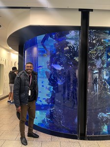 A man standing and smiling in front of a large, curved aquarium filled with colorful fish and coral. The aquarium has a blue hue from the water and lighting inside. The man is wearing a black jacket over a black shirt, brown pants, black shoes, and has a lanyard with an ID badge around his neck. The background includes a corridor with two people standing and walls with a textured design.