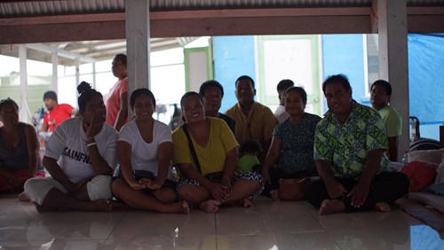 A group of Pacific Island community members sit barefoot on a shiny tiled floor inside a covered area with open walls. Most of them are smiling and facing the camera, dressed in casual clothing. The lighting is soft and natural, and the background shows a bright blue wall and some people standing or seated behind them, creating a relaxed and communal atmosphere.